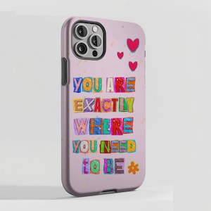 Può includere: Custodia per telefono lilla chiaro con la scritta "YOU ARE EXACTLY WHERE YOU NEED TO BE" in lettere colorate. La custodia ha un ritaglio per la fotocamera e tre cuori rossi. Un piccolo fiore giallo in basso.