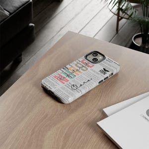 May include: A white phone case with a newspaper print design. The case has a colorful design with the words "Heroes Wear" and "RIN" printed on it. The case also has a black and white graphic of a bird with the letters "RN" printed on it.