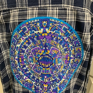 May include: A blue and white plaid flannel shirt with a circular patch on the back. The patch features a colorful Aztec-style design with yellow, purple, and blue details.