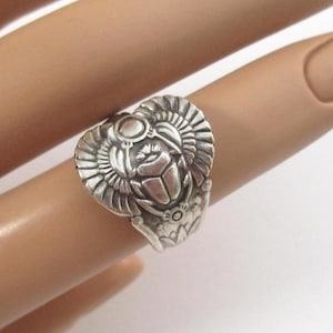 May include: A silver ring featuring a detailed scarab beetle design with prominent wings extending outward. The metal surface has a polished finish with darkened recessed areas that highlight the intricate carvings of the insect and its wings.