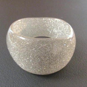 May include: A clear, glitter-infused bangle bracelet. The bracelet has a rounded, square shape and is filled with silver glitter, giving it a sparkling appearance. The bracelet is a fashion accessory.