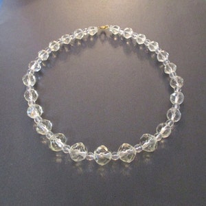 May include: A clear, faceted crystal necklace with a gold-colored clasp. The necklace is composed of multiple clear, round, and faceted beads. The beads are strung together to form a circular shape.