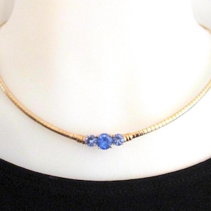 May include: A gold-toned necklace with a delicate chain and three blue gemstones. The gemstones are set in a row at the center of the necklace. The necklace is displayed on a white surface against a black background.
