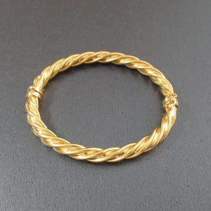 May include: A gold-toned bangle bracelet featuring a continuous twisted rope design. The circular bracelet has a polished, reflective surface and includes a small, integrated hinged clasp mechanism on one side to allow for opening and closing.