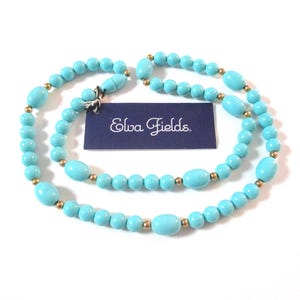 May include: A turquoise beaded necklace with gold-colored accents. The necklace features a mix of round and oval beads. A small tag with the text "Elva Fields" is attached to the necklace.