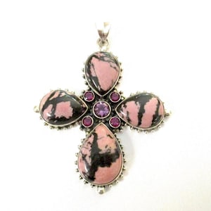 May include: A silver pendant in a cross shape, featuring large teardrop-shaped stones in pink and black, surrounded by smaller purple and red gemstones. The pendant has a detailed, ornate design.