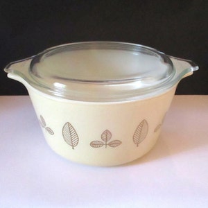 May include: A vintage, cream-colored Pyrex casserole dish with a clear glass lid. The dish features a brown leaf pattern around the exterior. The dish has two handles and is designed for oven use. A classic piece of kitchenware.
