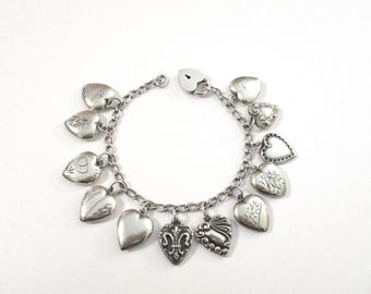 May include: Silver charm bracelet featuring heart-shaped charms. The bracelet includes a variety of heart designs, some with engraved patterns and others smooth. A heart-shaped padlock clasp secures the bracelet. The charms are suspended from a chain.