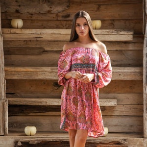 Puede incluir: Vestido rosa con hombros descubiertos y estampado de cachemira, con mangas largas y dobladillo corto. El vestido se combina con botines de ante marrones. El fondo es una estructura de madera.
