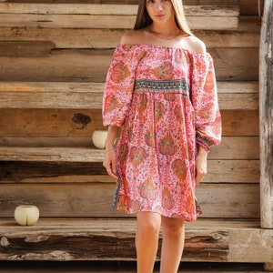 Puede incluir: Vestido rosa con hombros descubiertos con estampado de cachemira y mangas largas. El vestido tiene cintura entallada y se combina con botines marrones. El fondo es una estructura de madera.