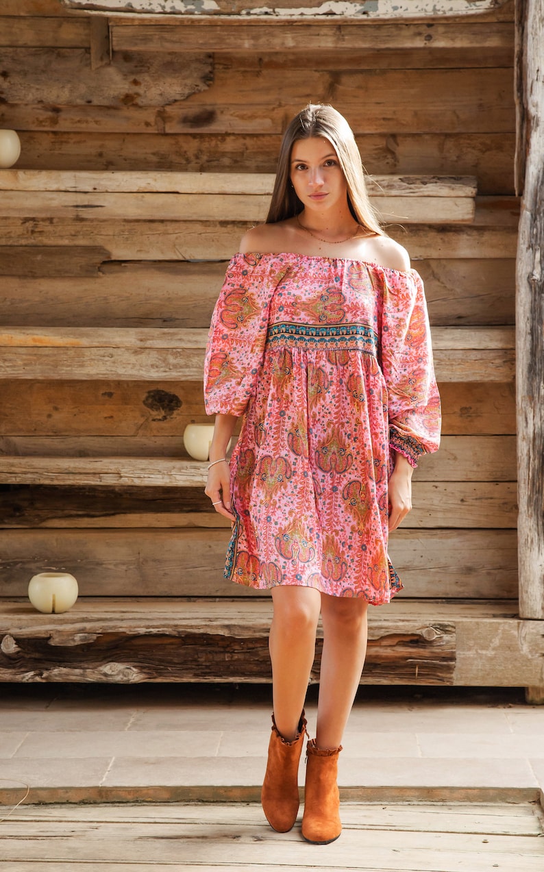 Puede incluir: Vestido rosa con hombros descubiertos con estampado de cachemira y mangas largas. El vestido tiene cintura entallada y se combina con botines marrones. El fondo es una estructura de madera.