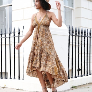May include: A woman wearing a halter-style maxi dress with a paisley pattern in shades of brown and gold. She is also wearing a straw hat and brown heeled sandals. The dress has a tiered skirt and a high-low hemline.