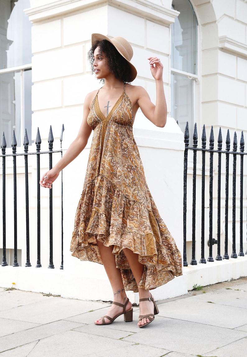 May include: A woman wearing a halter-style maxi dress with a paisley pattern in shades of brown and gold. She is also wearing a straw hat and brown heeled sandals. The dress has a tiered skirt and a high-low hemline.