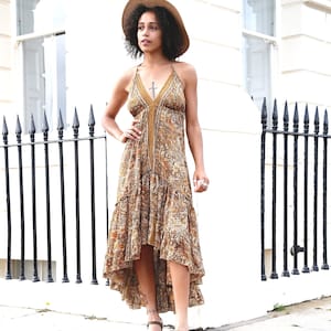 May include: A halter-neck maxi dress with a paisley pattern in shades of brown, tan, and gold. The dress has a high-low hemline and is paired with brown heeled sandals and a straw hat. The dress features a cross pendant.