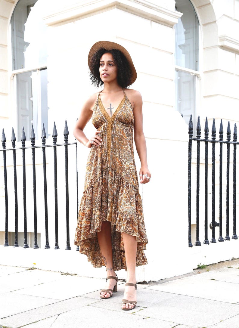 May include: A halter-neck maxi dress with a paisley pattern in shades of brown, tan, and gold. The dress has a high-low hemline and is paired with brown heeled sandals and a straw hat. The dress features a cross pendant.