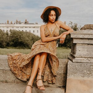 May include: A woman wearing a halter-neck maxi dress with a paisley pattern in shades of brown and gold, paired with brown heeled sandals and a wide-brimmed hat. The dress has a flowing skirt. The woman is sitting on a stone wall.