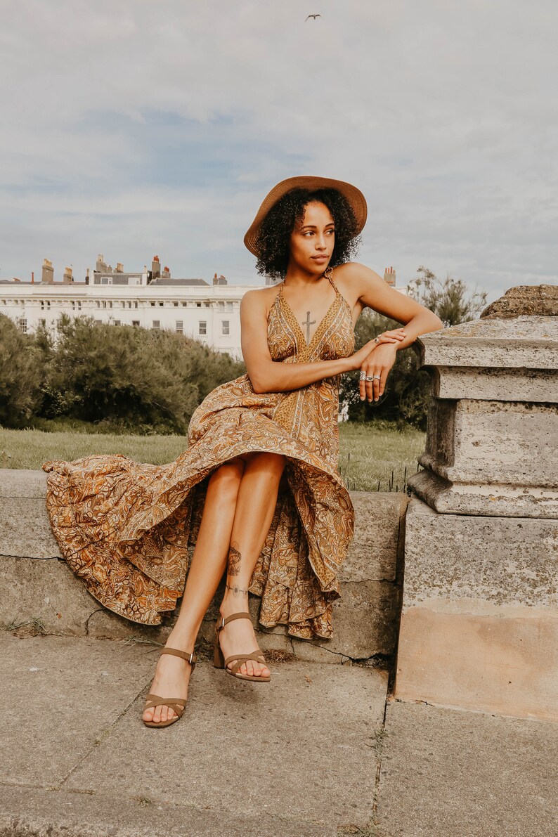 May include: A woman wearing a halter-neck maxi dress with a paisley pattern in shades of brown and gold, paired with brown heeled sandals and a wide-brimmed hat. The dress has a flowing skirt. The woman is sitting on a stone wall.