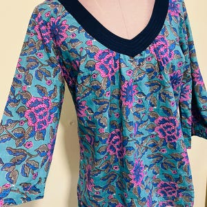 May include: A teal tunic top with a V-neck and three-quarter sleeves. The fabric is patterned with large pink and blue floral designs and gray leaves. The neckline is trimmed with a dark blue fabric.