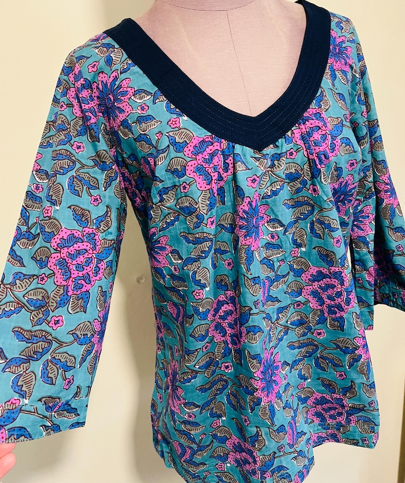 May include: A teal tunic top with a V-neck and three-quarter sleeves. The fabric is patterned with large pink and blue floral designs and gray leaves. The neckline is trimmed with a dark blue fabric.