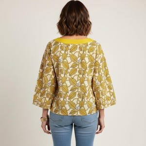 May include: A yellow and cream-colored blouse with a leaf pattern and a yellow neckline. The blouse has three-quarter length sleeves and is paired with blue jeans. The back view of the blouse is shown.