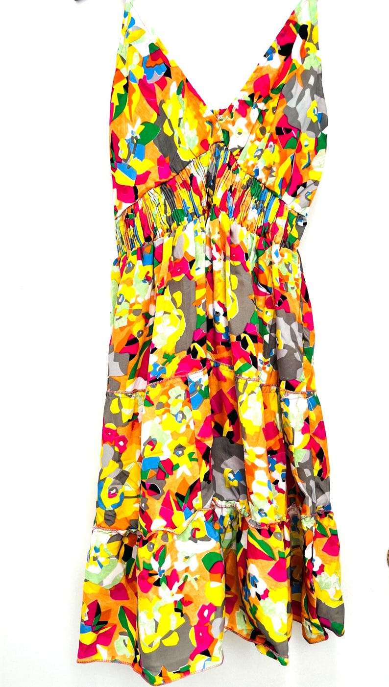 May include: A colorful summer dress with a V-neck and spaghetti straps. The dress features a vibrant abstract pattern in shades of yellow, orange, pink, blue, and gray. The dress has a tiered skirt and elastic waist.