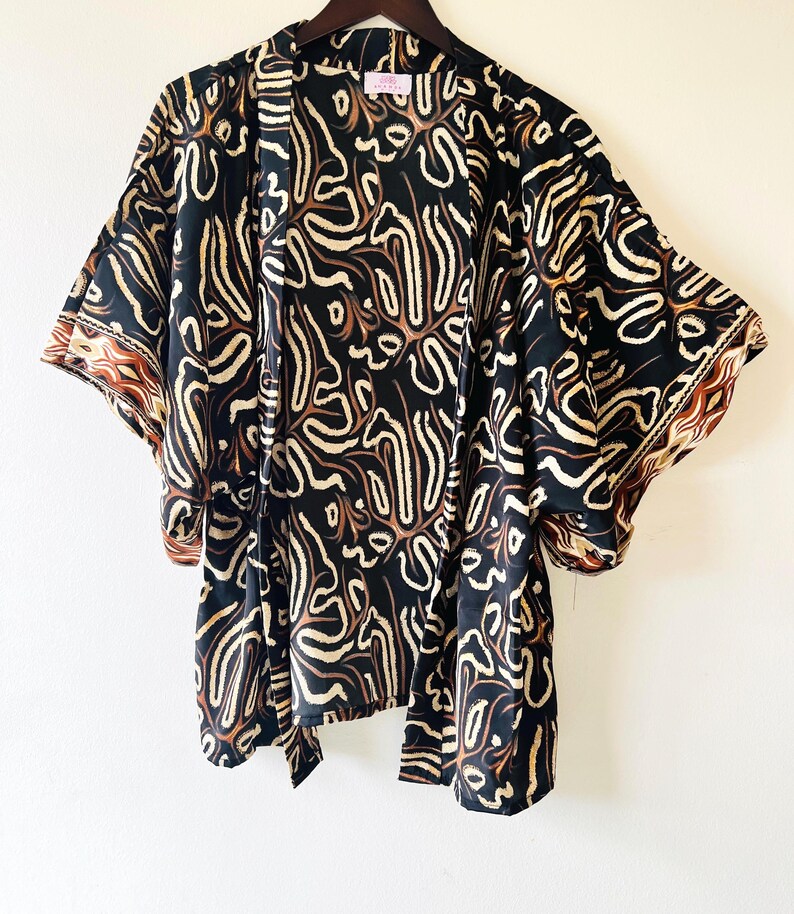 May include: Black kimono with a cream and brown abstract pattern. The kimono has wide sleeves and a tie closure. The edges of the sleeves have a brown and cream patterned border. The garment is hanging on a wooden hanger.