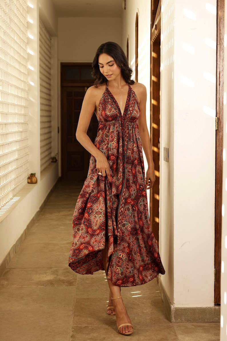 May include: A flowing, halter-neck dress with a paisley pattern in shades of red, brown, and teal. The dress features a deep V-neckline and a long, flared skirt. The model is wearing the dress in a well-lit hallway.