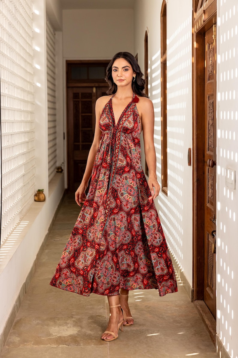 May include: A halter-neck midi dress with a paisley pattern in shades of red, pink, and blue. The dress features a deep V-neckline and a flowing skirt. The model is wearing nude-colored sandals.