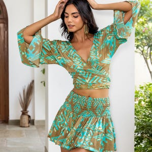 Op de afbeelding: Een tweedelige outfit met een wikkel top en een minirok. De set heeft een bruin en turquoise abstract patroon. De top heeft wijde mouwen en een V-hals. De rok heeft een gerimpelde tailleband en een gelaagd ontwerp.