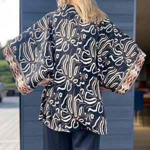 May include: A black kimono-style top with a cream and brown abstract pattern. The sleeves have a decorative border with geometric shapes in shades of brown and cream. The top is made of a flowing fabric.