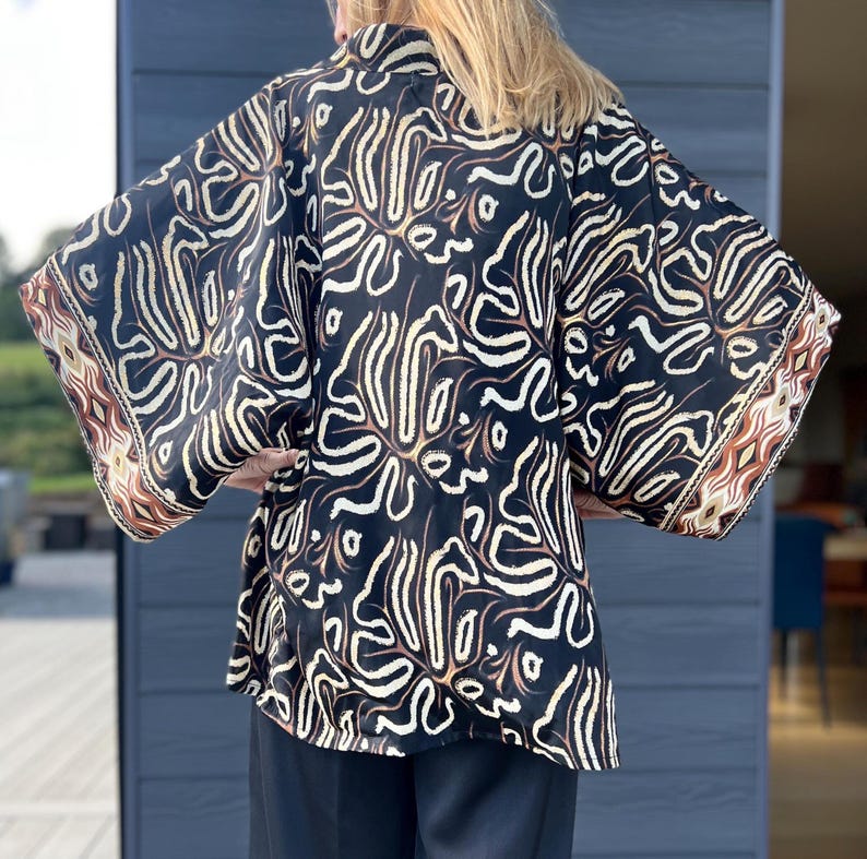 May include: A black kimono-style top with a cream and brown abstract pattern. The sleeves have a decorative border with geometric shapes in shades of brown and cream. The top is made of a flowing fabric.