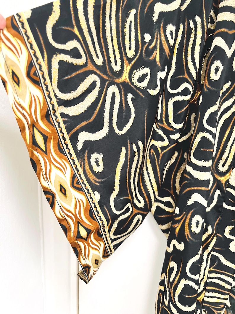 May include: A black garment with a cream and brown abstract pattern. The sleeve edge features a decorative border with geometric shapes in shades of brown and cream. The fabric appears to be lightweight and flowing.
