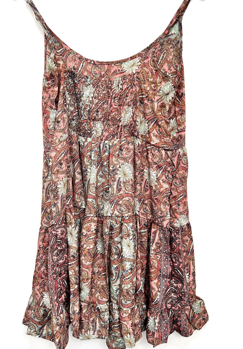 May include: A sleeveless dress with a paisley pattern in shades of pink, brown, and white. The dress features a tiered design with a gathered bodice and spaghetti straps. The fabric has a floral and paisley print.