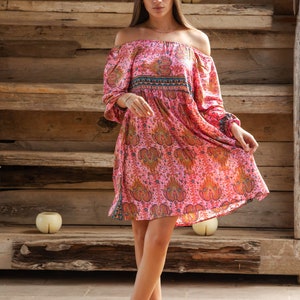 Puede incluir: Vestido rosa con hombros descubiertos y estampado de cachemira. El vestido tiene mangas largas y una falda fluida. La modelo lleva botines marrones. El fondo es una estructura de madera.