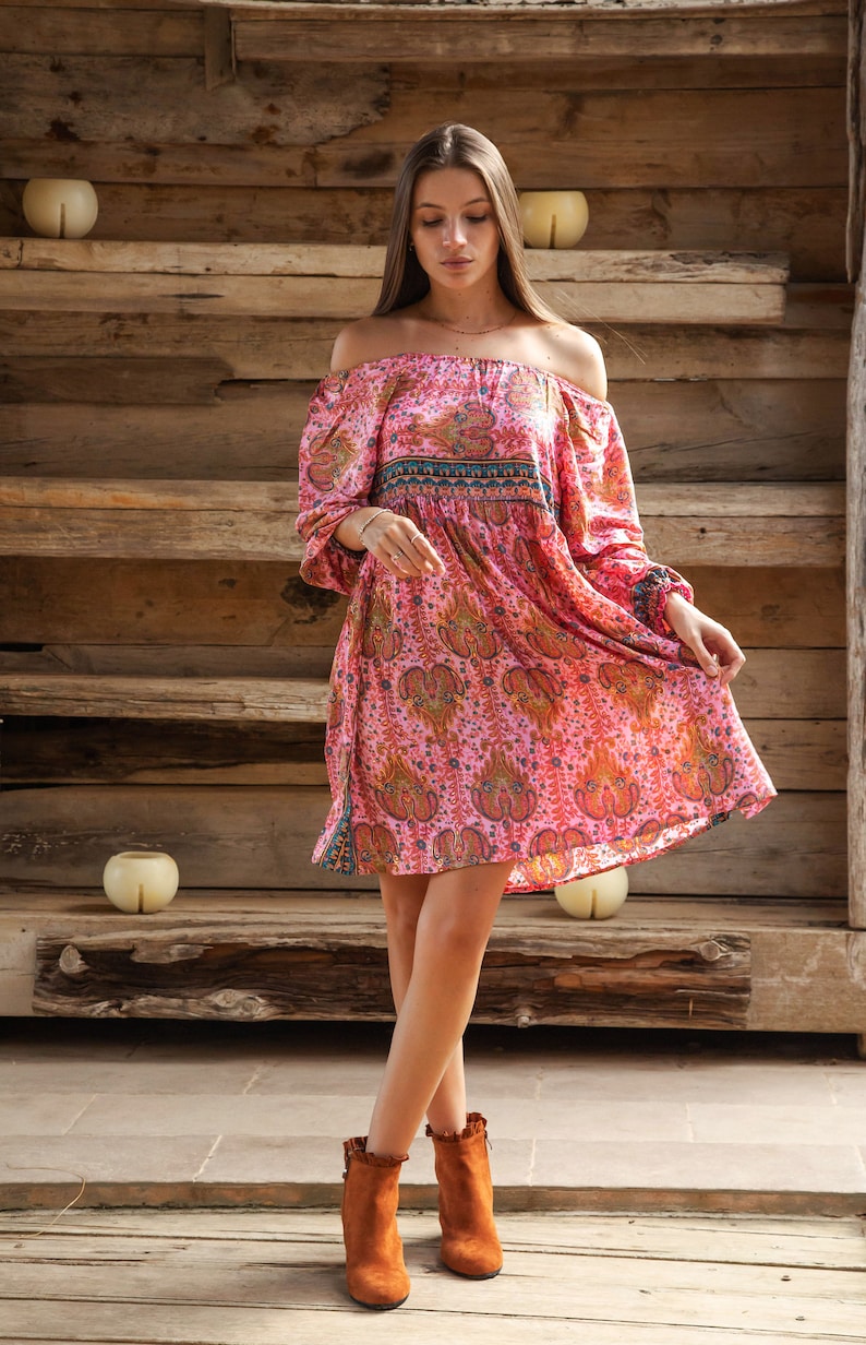 Puede incluir: Vestido rosa con hombros descubiertos y estampado de cachemira. El vestido tiene mangas largas y una falda fluida. La modelo lleva botines marrones. El fondo es una estructura de madera.