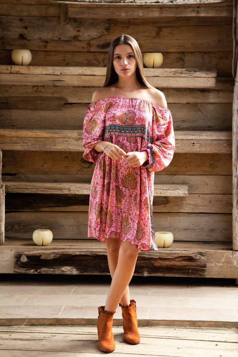 Puede incluir: Vestido rosa con hombros descubiertos con estampado de cachemira y mangas largas. El vestido tiene una banda decorativa azul y verde en el pecho. La modelo lleva botines marrones.