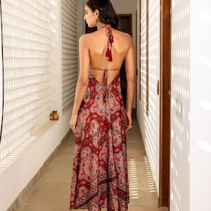 May include: A red paisley print halter maxi dress with a low back and tassel details. The dress features a flowing skirt and is worn with nude heels. The model is standing in a hallway with white walls and natural light.