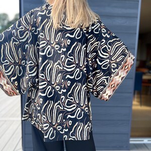 May include: A black kimono-style top with a cream and brown abstract pattern. The sleeves have a brown and cream geometric border. The top is paired with black pants. The model's blonde hair is visible.