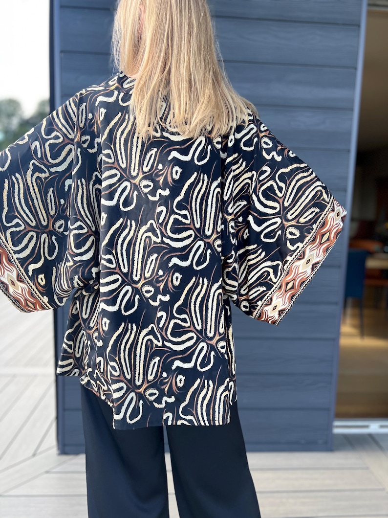 May include: A black kimono-style top with a cream and brown abstract pattern. The sleeves have a brown and cream geometric border. The top is paired with black pants. The model's blonde hair is visible.