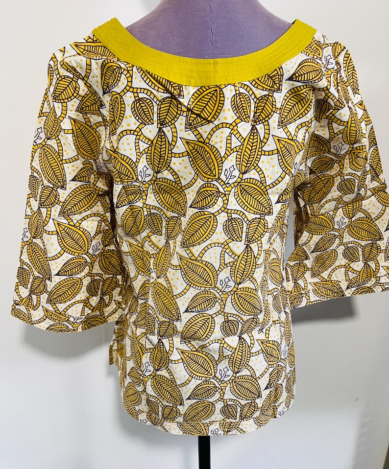 May include: A yellow and white patterned top with a leaf design. The top has three-quarter sleeves and a yellow neckline. The fabric is printed with a repeating pattern of yellow leaves and vines on a white background.