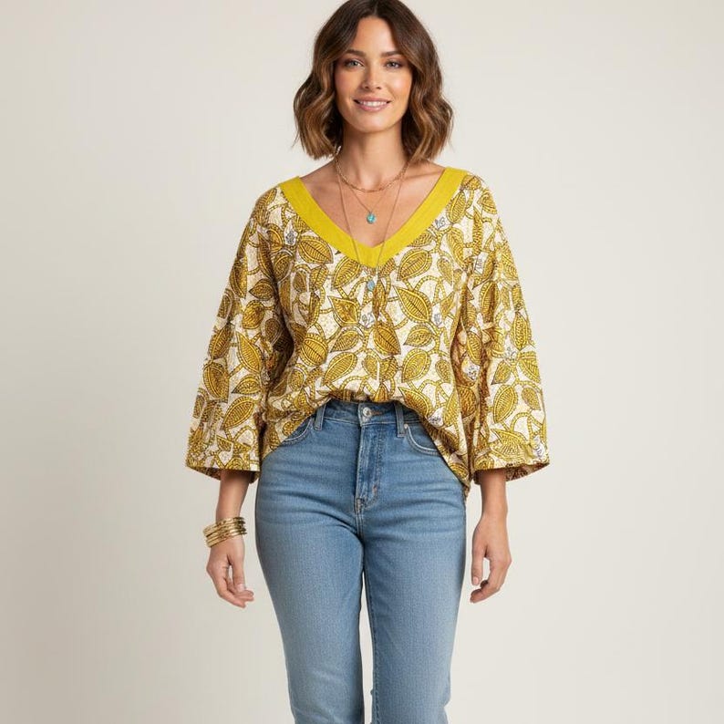 May include: A yellow V-neck blouse with a leaf pattern and three-quarter sleeves. The blouse has a yellow trim around the neckline. The model is wearing blue jeans and gold bracelets.