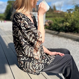 May include: A woman wearing a black and brown patterned top and black pants sits outdoors, holding a mug. The top features a V-neck and a unique abstract design. The background includes a wooden deck and a sunny sky.