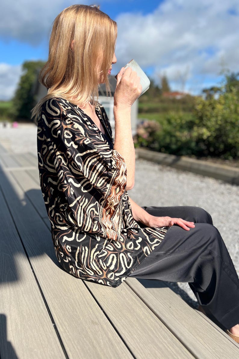 May include: A woman wearing a black and brown patterned top and black pants sits outdoors, holding a mug. The top features a V-neck and a unique abstract design. The background includes a wooden deck and a sunny sky.