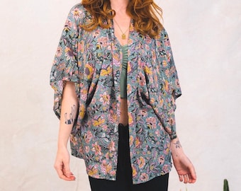 Peut inclure: Une veste kimono ouverte et ample, dotée d'un fond bleu sarcelle atténué avec un motif floral dense et multicolore dans des tons de jaune, rose et blanc. Le vêtement présente des manches larges arrivant aux coudes et une silhouette drapée.
