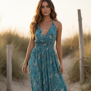 May include: A teal paisley maxi dress with a halter neck and a V-neckline. The dress has a flowing skirt. The model is wearing gold jewellery. The background features sand and tall grass.
