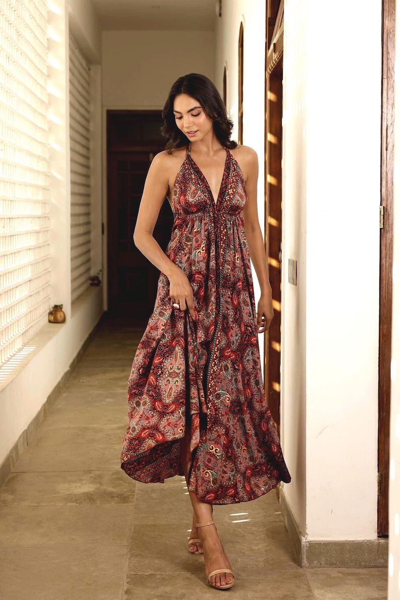 May include: A flowing, halter-neck dress in a paisley print featuring shades of red, brown, and teal. The dress has a V-neckline and a long, midi-length skirt. The model is wearing the dress in a well-lit hallway.