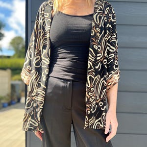 May include: A black kimono-style jacket with a cream and brown abstract pattern. The jacket is open, revealing a black tank top and black pants. The outfit is worn outdoors.