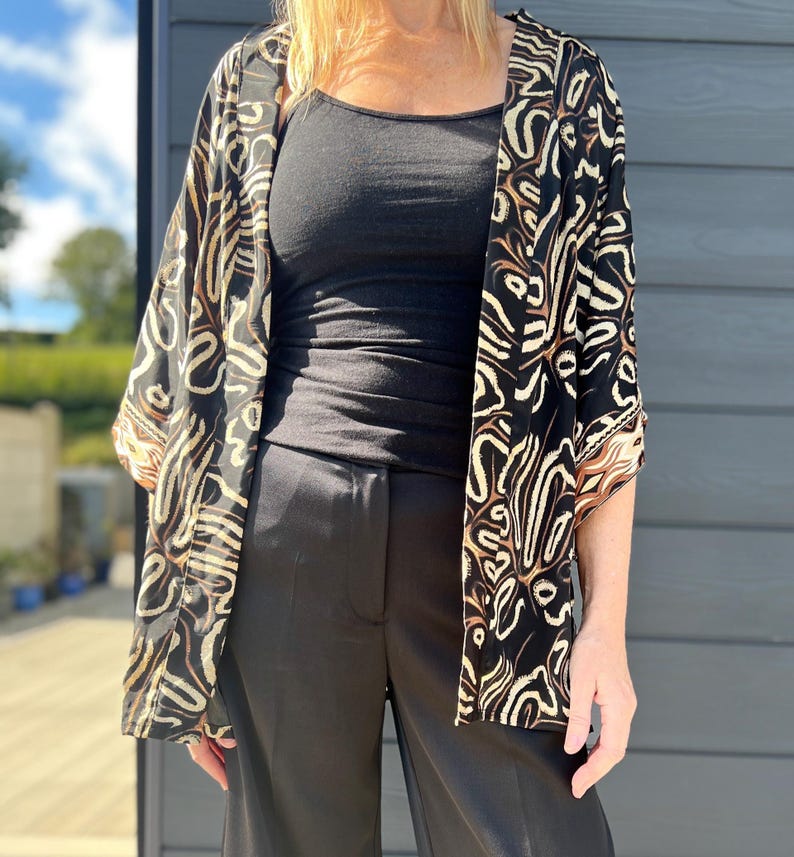 May include: A black kimono-style jacket with a cream and brown abstract pattern. The jacket is open, revealing a black tank top and black pants. The outfit is worn outdoors.