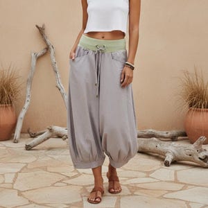 Puede incluir: Pantalones harén grises con cintura verde claro y cordón. Los pantalones tienen bolsillos y una forma abombada en los tobillos. La modelo lleva sandalias marrones y un top corto blanco. El fondo es de color neutro.