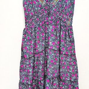 May include: A sleeveless, tiered dress with a V-neckline. The dress features a dark blue background with a floral pattern in pink and light blue. The neckline and hem are trimmed with a decorative border.